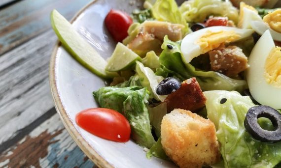 A close-up of a Caesar salad featuring fresh lettuce, eggs, croutons, and olives on a rustic table.