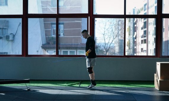 Young man exercising with a skipping rope in an urban gym with large windows and city view.